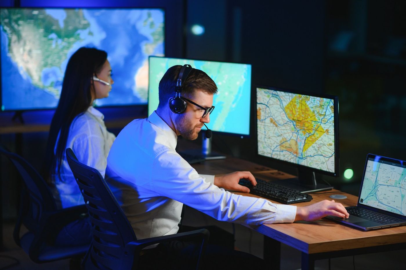 Two telecommunicators in headsets work at mapping displays in a dimly lit communications center.