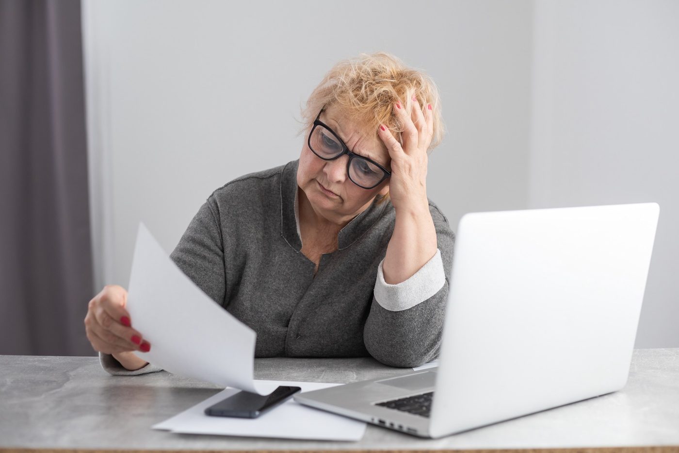 A woman at her kitchen table reads a bill with a worried look, her laptop open beside her.