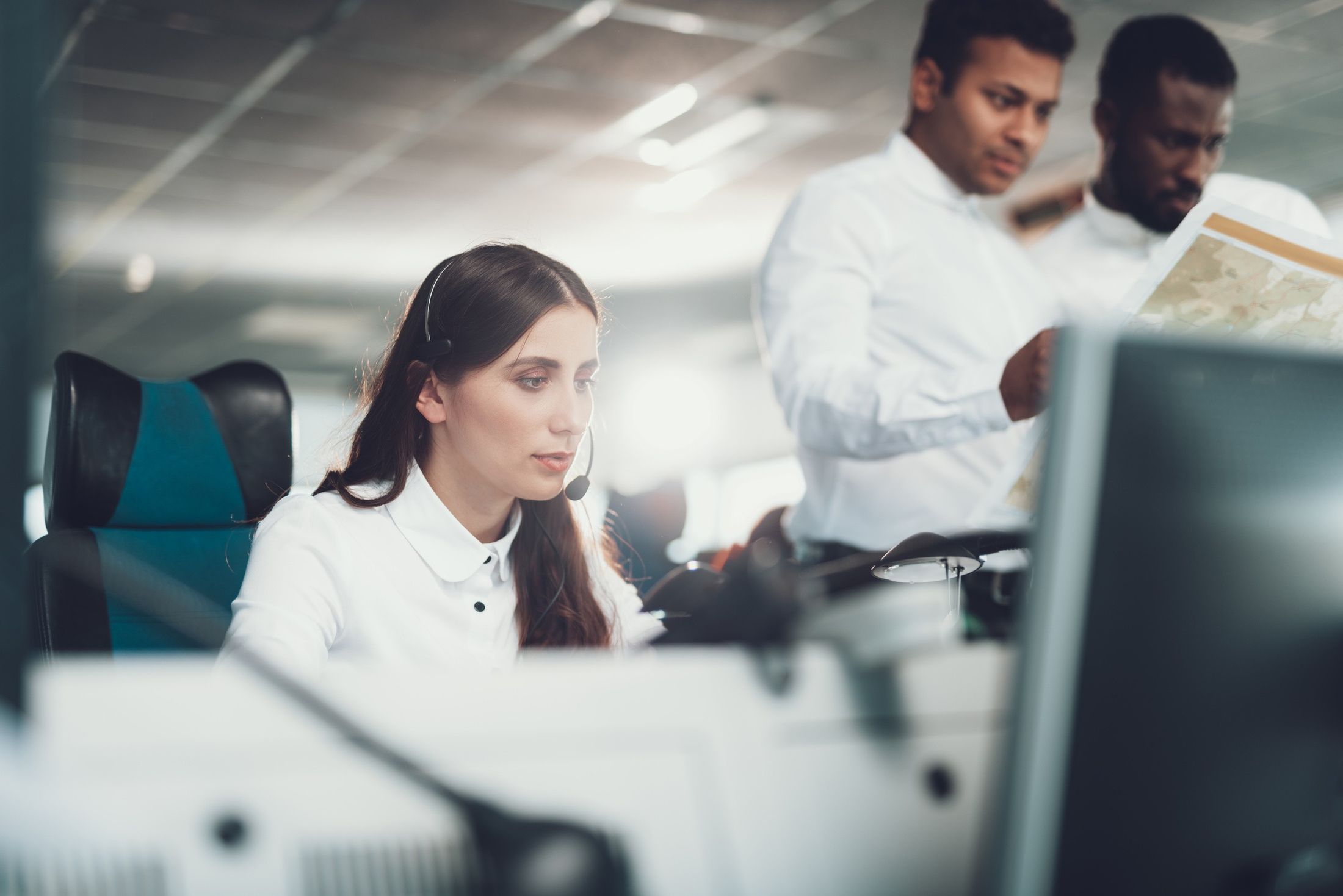 A dispatcher wearing a headset works at her console while two colleagues review a map behind her.